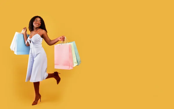 Full length of carefree black woman in dress running with bright shopping bags over orange background, free space — стоковое фото