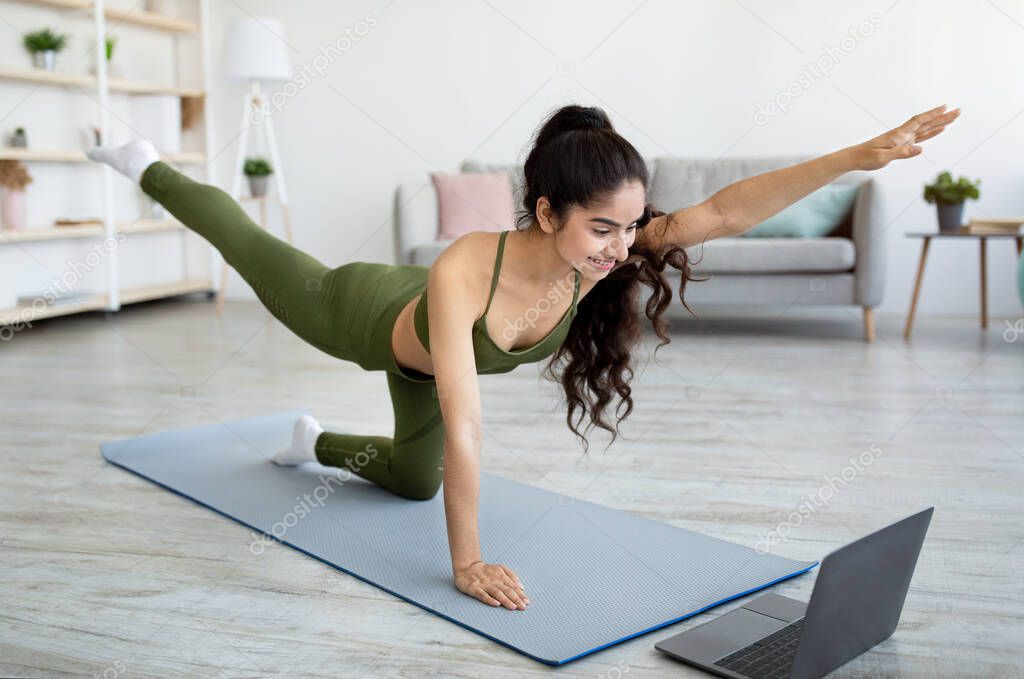 Online sports during covid lockdown. Millennial Indian woman exercising on mat, watching sports video on laptop at home