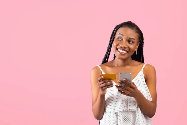Milenial African American Lady memegang kartu kredit dan ponsel, melihat ruang kosong di latar belakang studio merah muda — Stok Foto