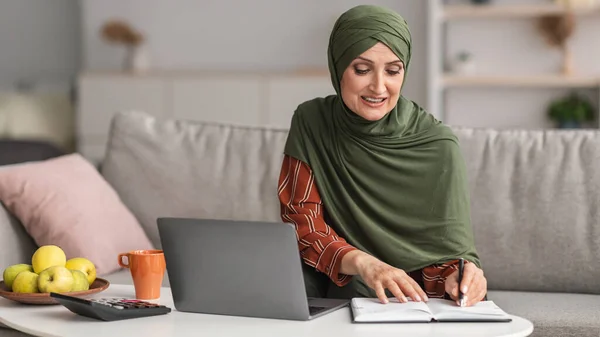 Счастливая зрелая исламская леди с помощью ноутбука принимая заметки дома — стоковое фото
