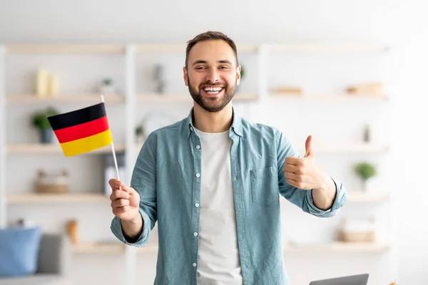 Felice uomo caucasico mostrando pollice in su e la bandiera della Germania, in posa e sorridente alla fotocamera al chiuso — Foto Stock