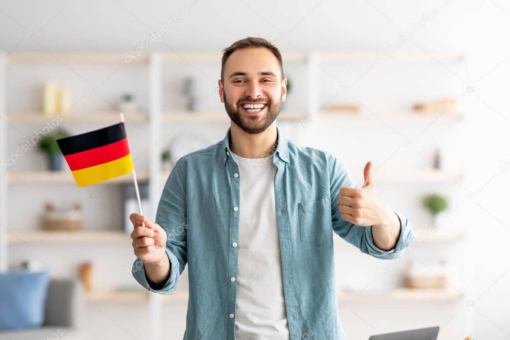 Happy Caucasian man showing thumb up and flag of Germany, posing and smiling at camera indoors