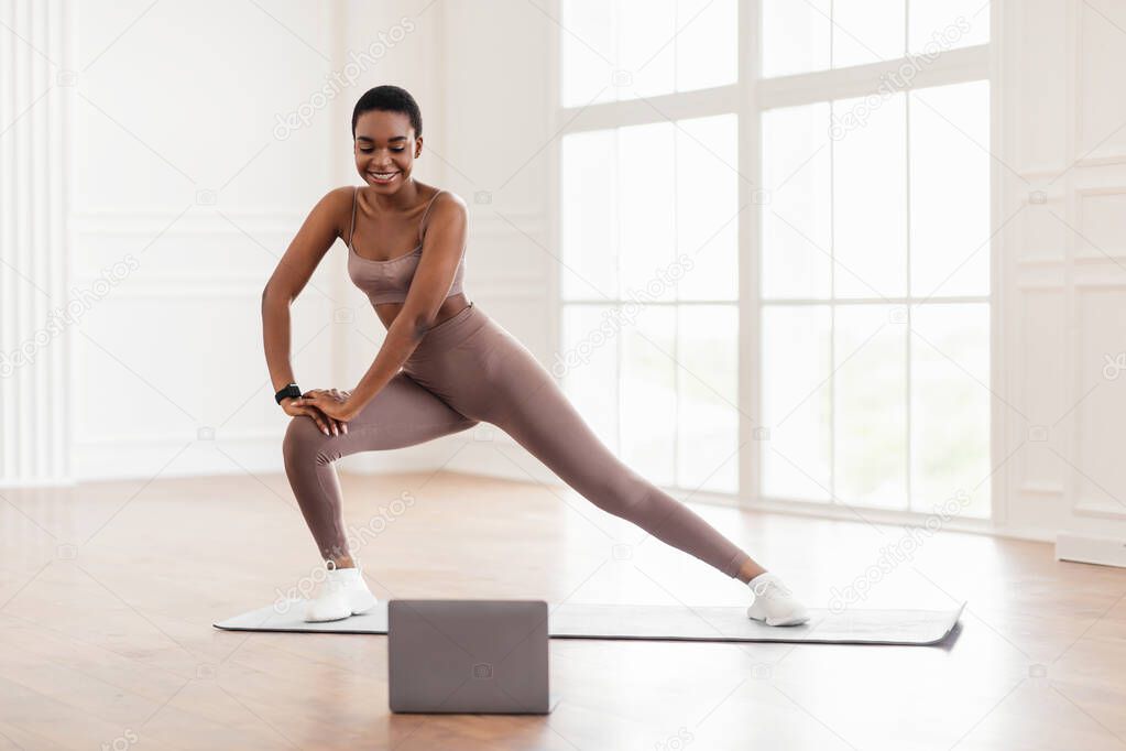 Young black woman doing side lunges on mat using laptop