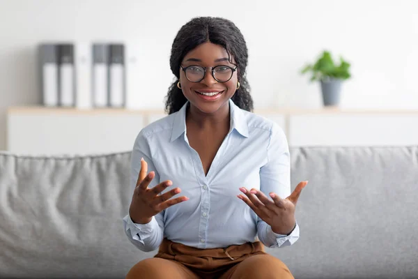 Porträtt av leende ung svart psykolog gestikulerar och ler mot kameran, sitter på bekväm soffa på modernt kontor — Stockfoto