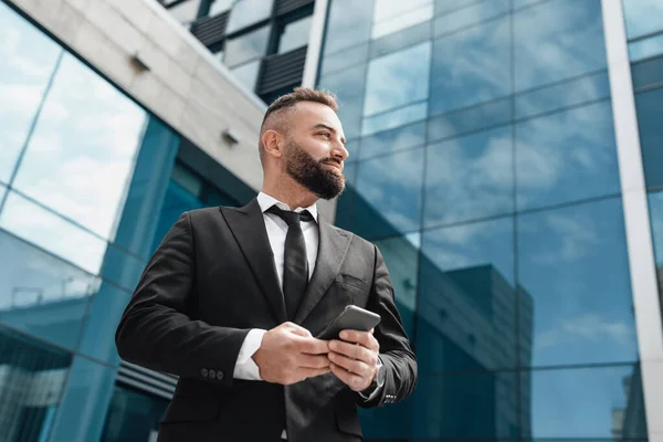 Empresario seguro en traje negro que sostiene el teléfono inteligente, de pie contra el centro de negocios de vidrio moderno, espacio libre — Foto de Stock