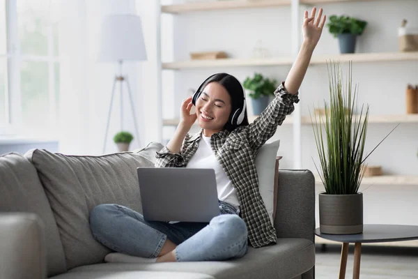 Ung asiatisk kvinna i trådlösa hörlurar njuter av musik, med hjälp av bärbar dator — Stockfoto