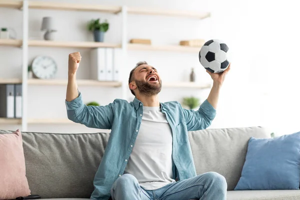 Συναισθηματικός τύπος βλέποντας ποδόσφαιρο στην τηλεόραση, σηκώνοντας μπάλα πάνω από το κεφάλι του και φωνάζοντας, γιορτάζοντας γκολ ή νίκη στο σπίτι — Φωτογραφία Αρχείου