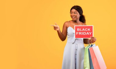 Elinde siyah alışveriş torbaları olan çekici bir zenci kadın. Elinde Kara Cuma tabelası var ve boş alanı işaret ediyor. Turuncu arka plan.