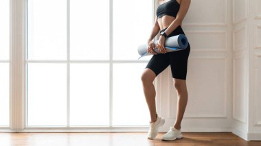 Pencere kenarında yoga minderi tutan genç bir kadın.