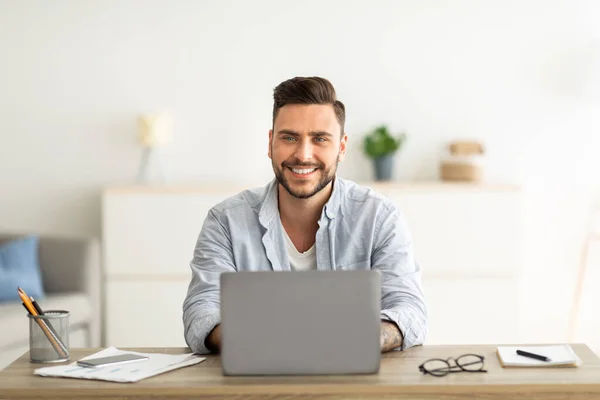 Libero professionista di successo. Uomo felice seduto sul posto di lavoro con computer portatile, sorridente e guardando la fotocamera — Foto Stock