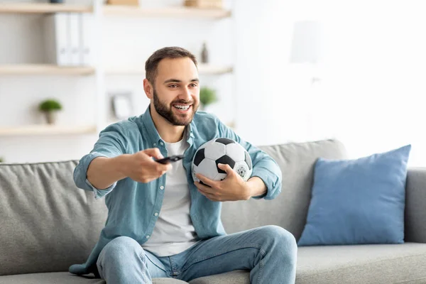 Glücklicher junger Kerl schaut Fußballspiel im Fernsehen und hält Fernbedienung und Ball zu Hause — Stockfoto
