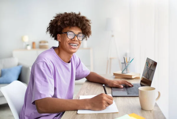 Conceito de e-learning. Retrato de adolescente afro-americano sorridente tendo aula na web sobre laptop, tomando notas em casa — Fotografia de Stock