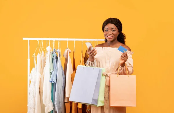 Happy zwarte vrouw met smartphone voor online winkelen, staan in de buurt van kledingrek met shopper tassen en credit card — Stockfoto