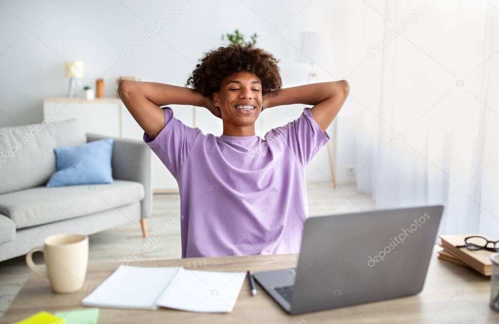 Cheerful black teen guy holding hands behind head, relaxing in front of laptop at home