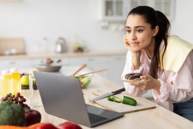 Dizüstü bilgisayar kullanan ve tatlı yiyen endişeli bir kadın.