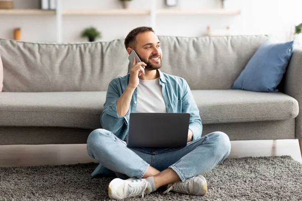 Full length of happy young guy using laptop and mobile phone στο σπίτι, έχοντας ανεξάρτητη εργασία — Φωτογραφία Αρχείου