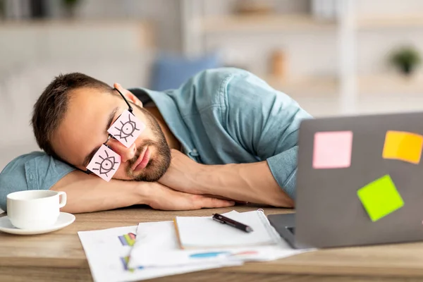 Ленивый непродуктивный молодой парень в смешных липких нотах с открытыми глазами на очках, спит на рабочем месте — стоковое фото