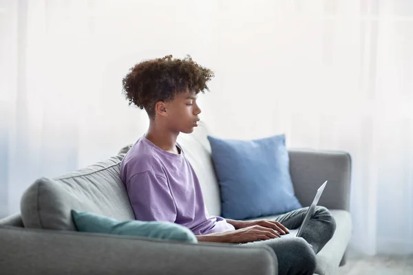 Η ιδέα της εξ αποστάσεως εκπαίδευσης. Μαύρος έφηβος που χρησιμοποιεί φορητό υπολογιστή για τις online σπουδές του στο σπίτι, πλαϊνή όψη — Φωτογραφία Αρχείου