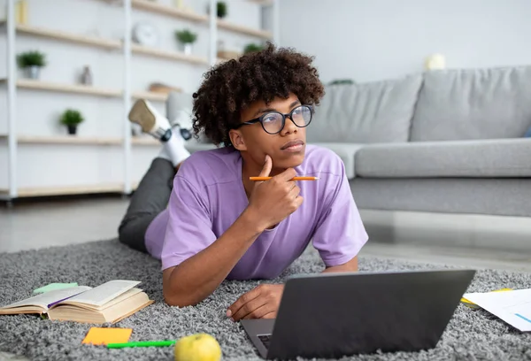 Tankeväckande svart tonåring kille som använder laptop för fjärrstudier, liggande på golvet hemma — Stockfoto