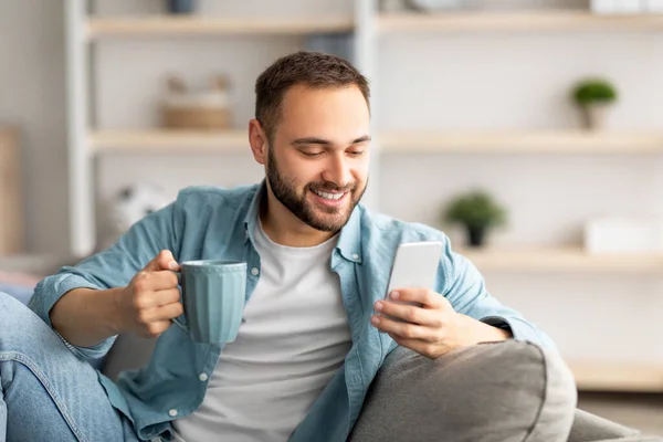 Pozitív fiatal srác kezében okostelefon és kávé zárt térben, csevegés barátaival online, e-mail küldése, böngészés internet — Stock Fotó