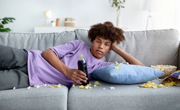 Verveeld zwart tiener liggend op bank met verspreid voedsel, kijken saaie show of film op tv, doden tijd thuis — Stockfoto