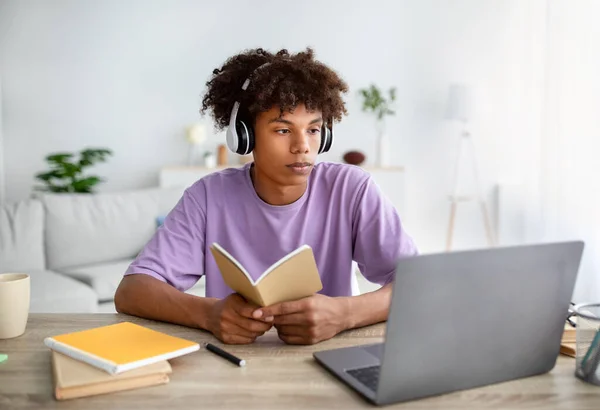 Inteligentny czarny nastolatek student w słuchawkach studiuje online przed laptopem, trzymając notatnik w domu — Zdjęcie stockowe