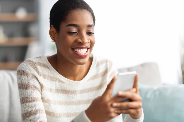 Alegre preto mulher usando smartphone enquanto relaxante no sofá em casa — Fotografia de Stock