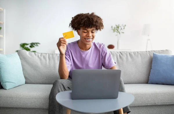 Concepto de compra remota. Adolescente negro positivo con el ordenador portátil y la tarjeta de crédito que ordena mercancías en línea del hogar —  Fotos de Stock