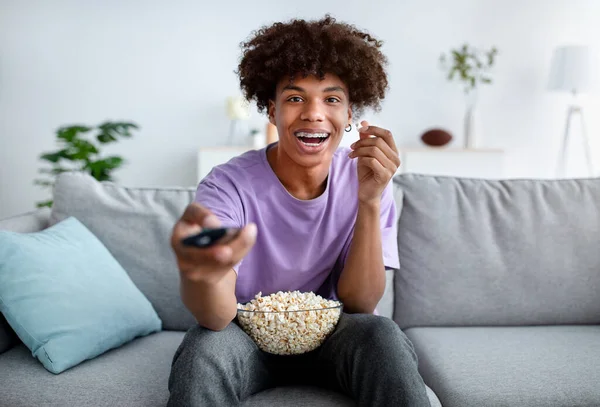 Divertissements à la maison. Adolescent afro-américain excité avec contrôleur TV appréciant le film et mangeant du pop-corn à l'intérieur — Photo