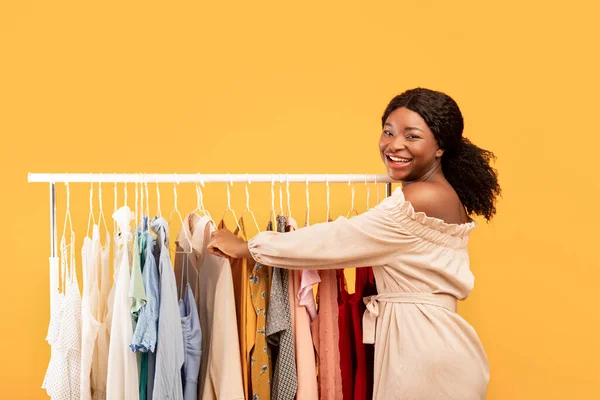 Seizoensgebonden winkelconcept. Zwarte vrouw kiezen kleding uit rek, volgens de nieuwste modetrends op oranje achtergrond — Stockfoto