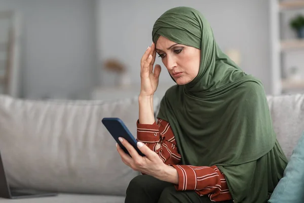 Frustrada musulmana dama sosteniendo el teléfono celular leyendo malas noticias en casa — Foto de Stock