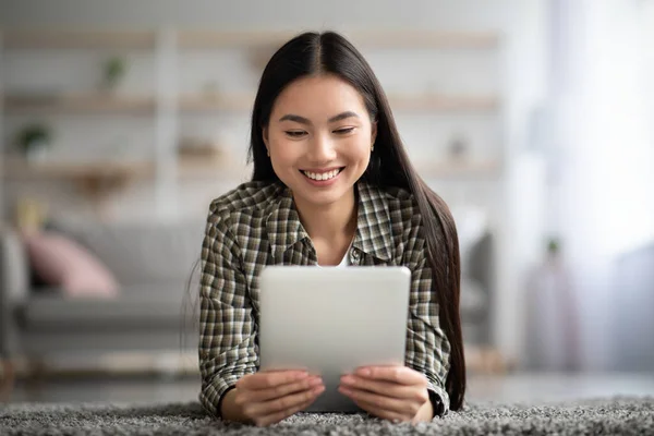 Vrolijke aziatische dame ligt op tapijt thuis, met behulp van pad — Stockfoto