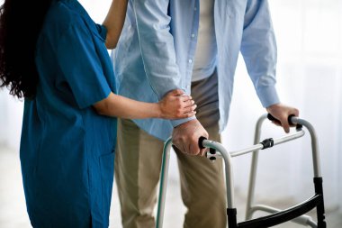 Yaşlı bir adamın Walker 'ı kullanması, genç bir hemşirenin huzurevinde ona destek olması ve yardım etmesi, yakın plan.