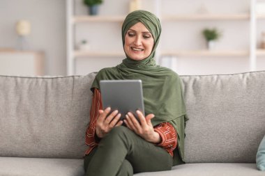 Evde oturan Orta yaşlı Müslüman kadın tablet bilgisayar kullanıyor.