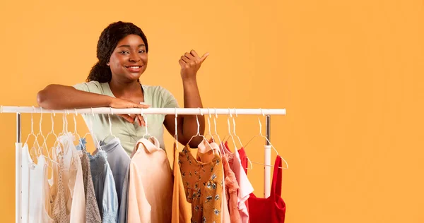 Retrato de jovem senhora negra em pé na loja de roupas com roupas elegantes no rack, apontando para o espaço vazio — Fotografia de Stock