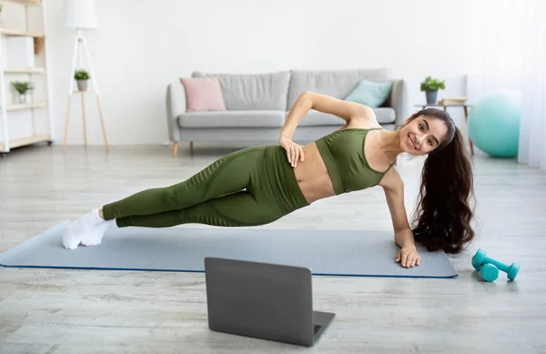 Online sporten tijdens gezelligheid. Fit Indiase vrouw doet oefeningen in de voorkant van de laptop, staan in de zijkant plank thuis — Stockfoto