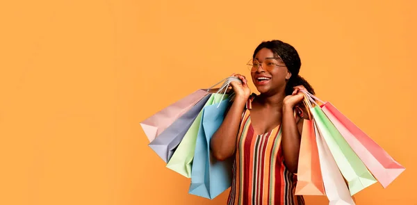 Atractiva joven mujer negra en ropa de verano sosteniendo bolsas de regalo, mirando el espacio en blanco sobre fondo de estudio naranja —  Fotos de Stock