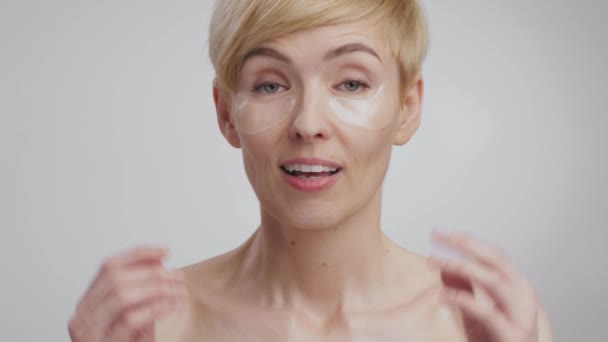 Emotional middle aged lady with collagen patches feeling amazed, raising brows and hands, grey studio background — Stock Video