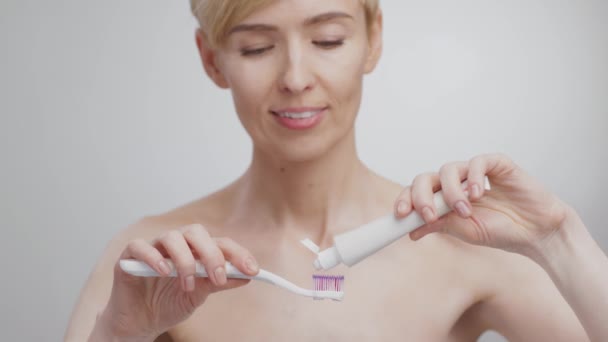 Mujer rubia de mediana edad aplicando pasta de dientes en el cepillo de dientes y sonriendo a la cámara, fondo gris estudio — Vídeo de stock
