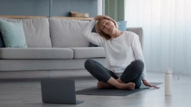 Yetişkin kadın dizüstü bilgisayarda egzersiz yapıyor. Boynunu esnetiyor. Evde oturuyor.