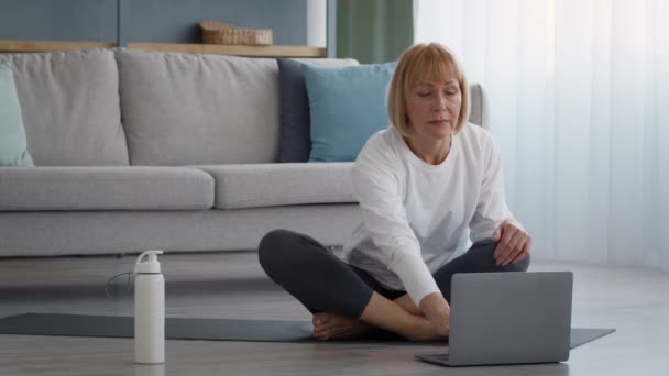 Sportliche Seniorin sitzt mit Laptop auf Gymnastikmatte — Stockvideo