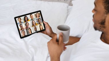 Yatakta dijital tabletli siyahi adam bir grup insanla telekonferans yapıyor.