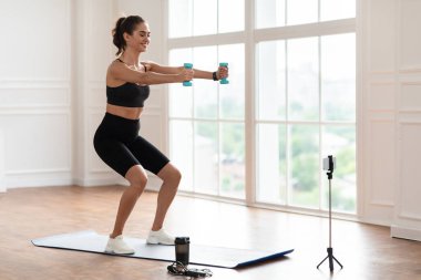 Akıllı telefonun önünde dambıllarla egzersiz yapan genç bir kadın.