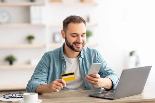 Ευτυχισμένος Καυκάσιος αγοράζοντας πράγματα online, χρησιμοποιώντας smartphone, laptop και πιστωτική κάρτα, απολαμβάνοντας τα ψώνια στο διαδίκτυο — Φωτογραφία Αρχείου