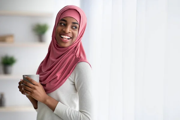 Felice nero musulmano donna in hijab bere caffè vicino finestra a casa — Foto Stock