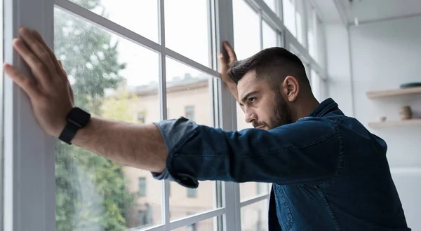 Hombre bajo cuarentena en casa, de pie frente a la ventana, con la mano en el vidrio, tristemente mirando hacia fuera. Depresión de bloqueo — Foto de Stock