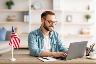 ABD bayrağı taşıyan mutlu genç adam ofis laptopuyla masa başında çalışıyor.