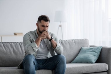 Düşünceli, genç, ciddi, yakışıklı bir adam evdeki kanepede oturur, düşünceler içinde kaybolur, problem çözmeyi düşünür.