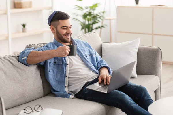 Smiling jewish man watching video on computer, drinking hot coffee — Stock Photo, Image
