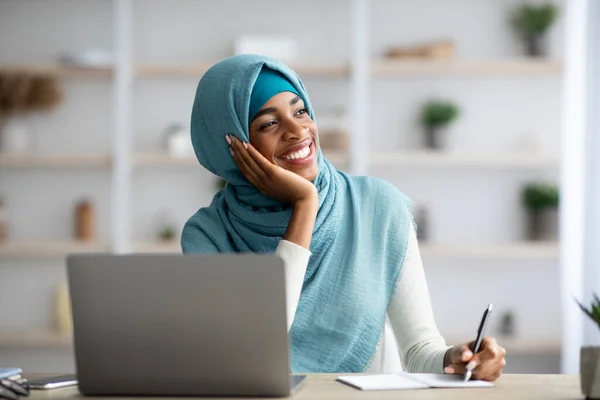 Szczęśliwy czarny muzułmanin pani siedzi przy biurku z laptopem i patrząc w bok — Zdjęcie stockowe